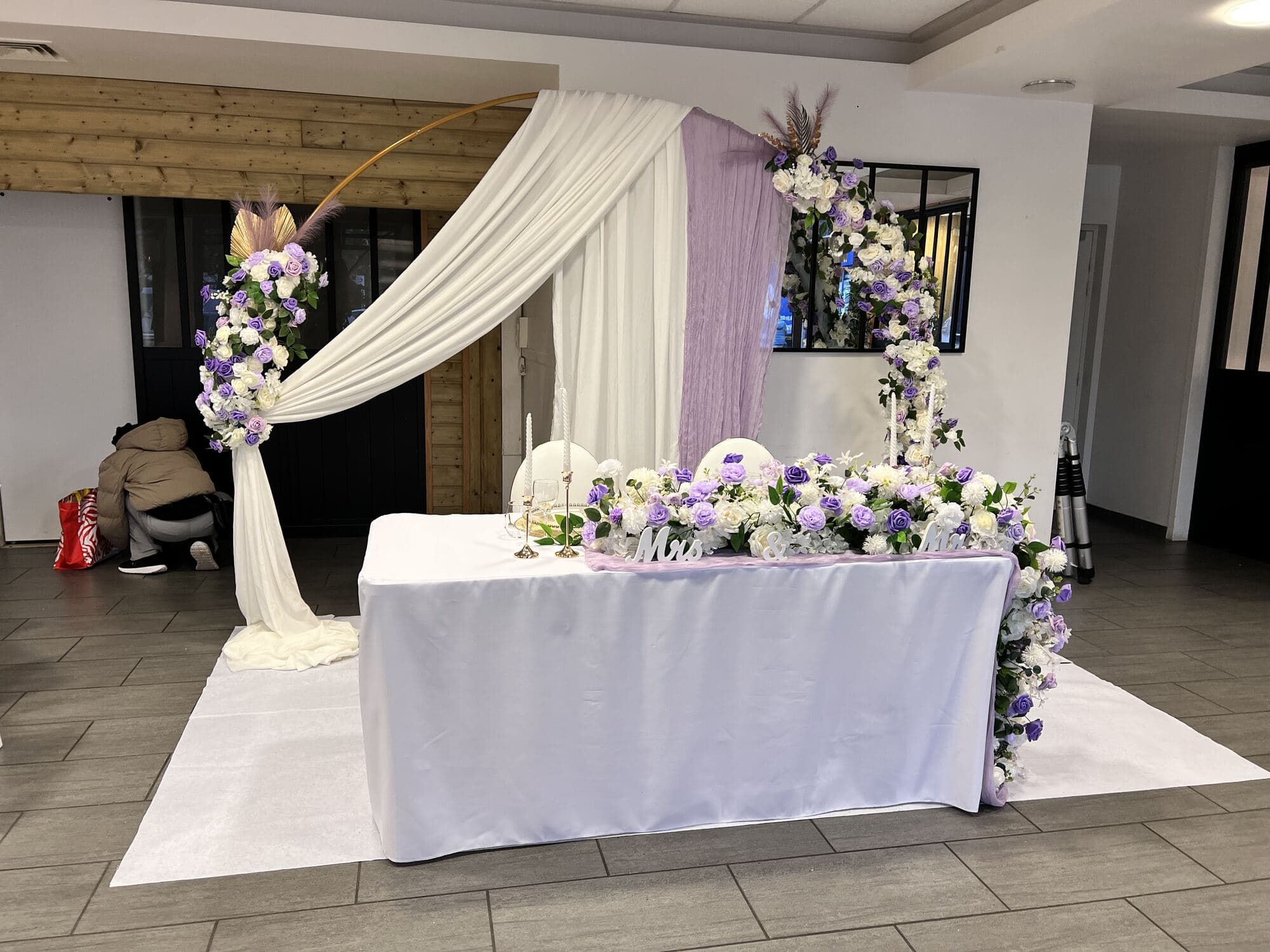 Coin des mariés lilas et doré : table d'honneur sous arche drapée et chemin de roses blanches — Salle du Pavillon, Épône